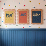 Three framed educational wall art prints hanging on a wall above blue wainscoting. The prints feature the words PLAY, LOUD, and DREAM with definitions, styled with colorful star decals on a white wall.