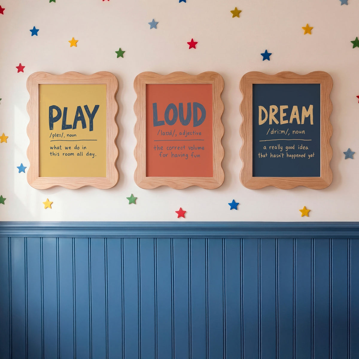 Three framed educational wall art prints hanging on a wall above blue wainscoting. The prints feature the words PLAY, LOUD, and DREAM with definitions, styled with colorful star decals on a white wall.
