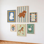Six framed whimsical art prints arranged in a gallery wall on a white wall above a light wood floor. The prints feature a white poodle on blue, a brown Highland cow on gray stripes, a blue watering can with tulips, a brown horse, and a white goose.