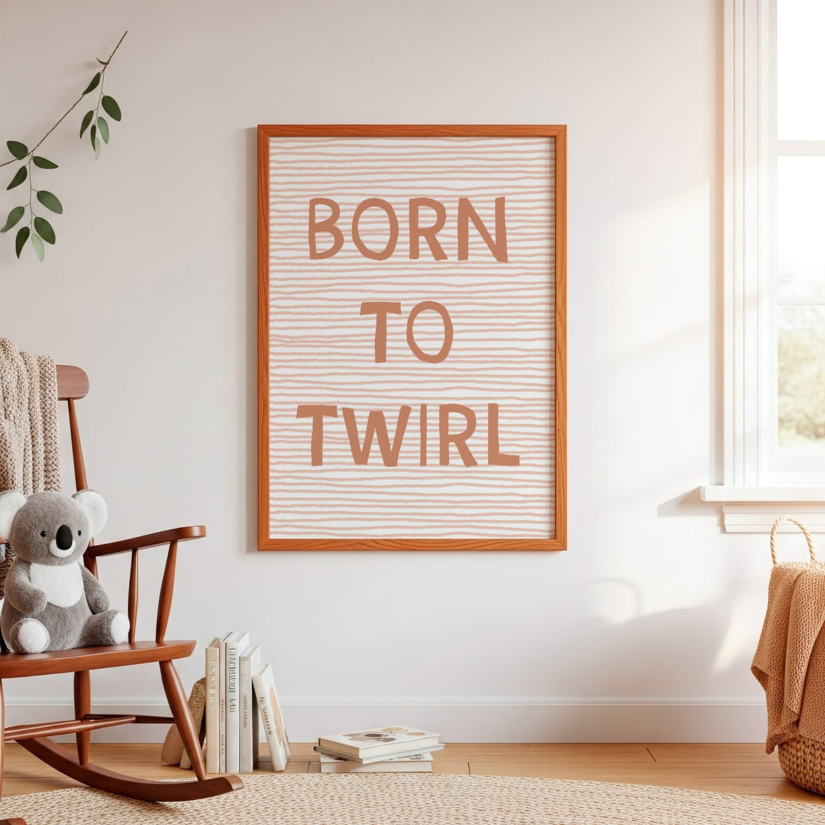 A framed nursery wall art print reading 'BORN TO TWIRL' in peach lettering on a white background with thin horizontal peach stripes, hanging on a white wall in a sunlit room with a wooden rocking chair, books, and a koala toy.