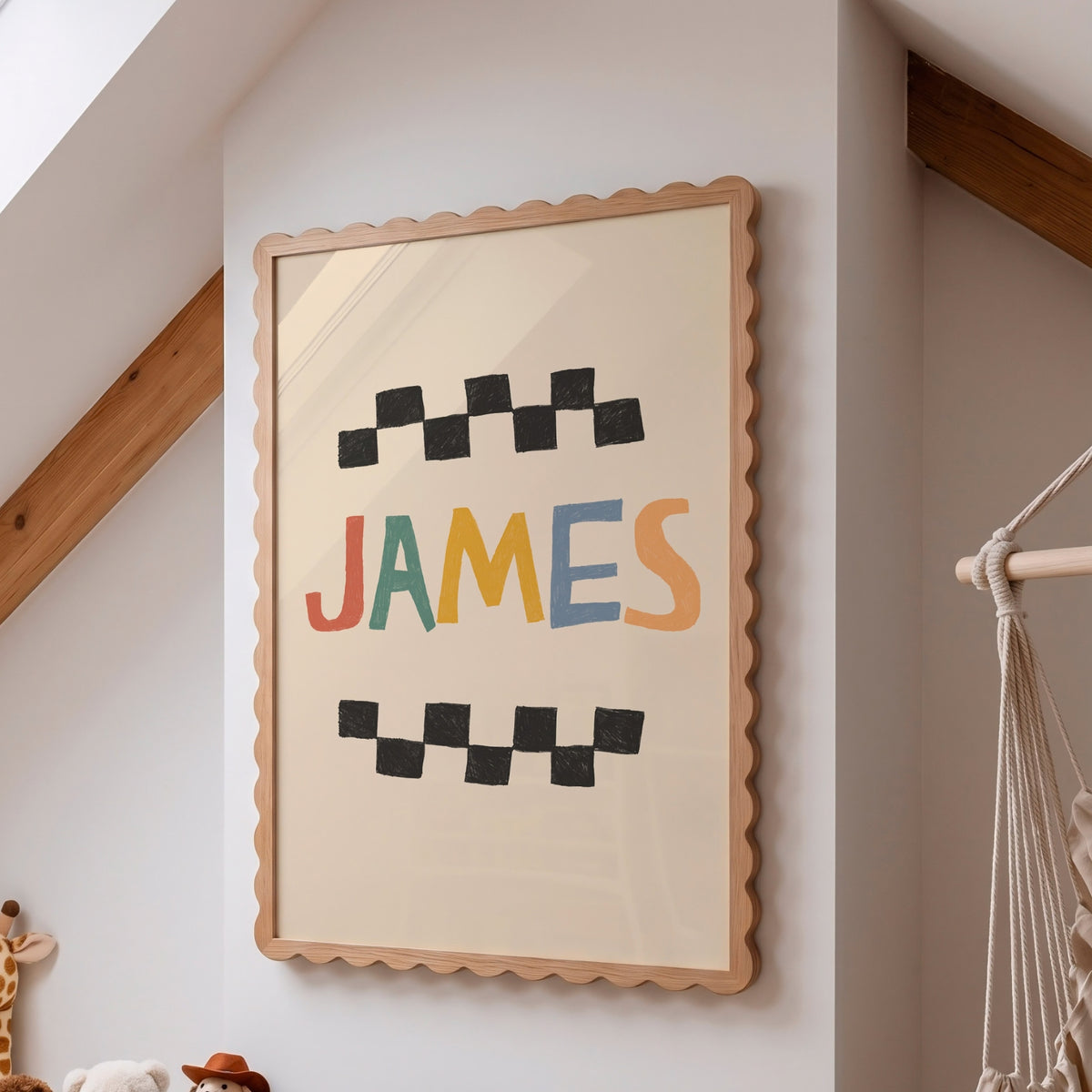 A framed personalized wall art print featuring the name 'JAMES' in colorful letters between black checkered patterns, hanging on a white wall in a room with wooden beams and a macrame hanging visible on the right.