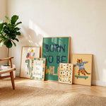 Five framed art prints with colorful designs are arranged on a light wood floor against a beige wall in a styled room setting with a potted plant and a chair.