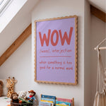 A framed educational wall art print featuring the word 'WOW' with phonetic spelling and a definition, styled in a child's attic bedroom next to a macrame hanger and toys.