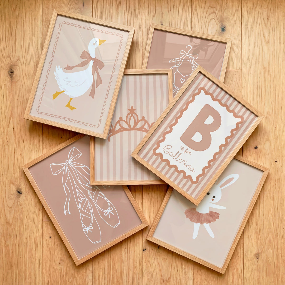Six framed nursery art prints with ballet and baby-themed illustrations are arranged in a flat-lay on a light wood floor. The prints feature a duck, a ballerina, a crown, ballet slippers, a bunny in a tutu, and a large letter B with the text 'is for Ballerina'.