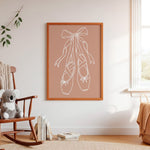 A framed wall art print featuring a white line drawing of ballet pointe shoes on a dusty pink background, styled in a warm, neutral nursery or children's room with a wooden rocking chair, plush toy, and window.