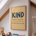 A framed educational wall poster with a scalloped wooden frame hangs on a white wall in a child's room. The poster features the word 'KIND' in large dark blue letters with its pronunciation '/kænd/, adjective' and the phrase 'the coolest thing you can be' written below.