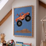 A framed children's wall art print featuring a cartoon orange monster truck jumping over a brown hill on a blue background, displayed on a white wall in a nursery or kids' room with toys and a hammock visible.