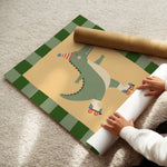A person is unrolling a large decorative floor mat or rug on a beige carpet. The mat features a green and white striped border and a central illustration of a green alligator wearing roller skates and a party hat on a pale yellow background.