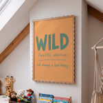 A framed wall art print with a scalloped wooden frame hangs on a white wall in a child's room. The print features the word 'WILD' in large teal letters with a dictionary-style definition underneath: '/wazld, adjective not always a bad thing'.