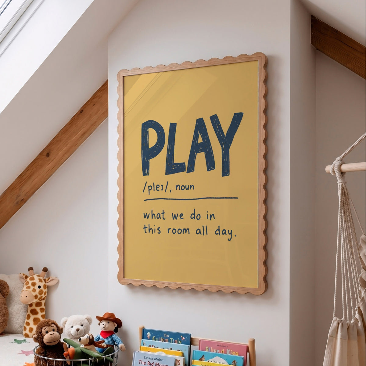 A framed children's wall poster with a scalloped light wood frame hangs on a white wall in a bright attic room. The poster has a mustard yellow background and reads “PLAY /ples/, noun” followed by the definition “what we do in this room all day.