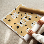 A person unrolls a large cookie-themed wall decal or poster on a beige carpet, with a brown paper roll nearby. The design shows a tan cookie with black polka dots and white text reading 'OK LET'S PLAY'.