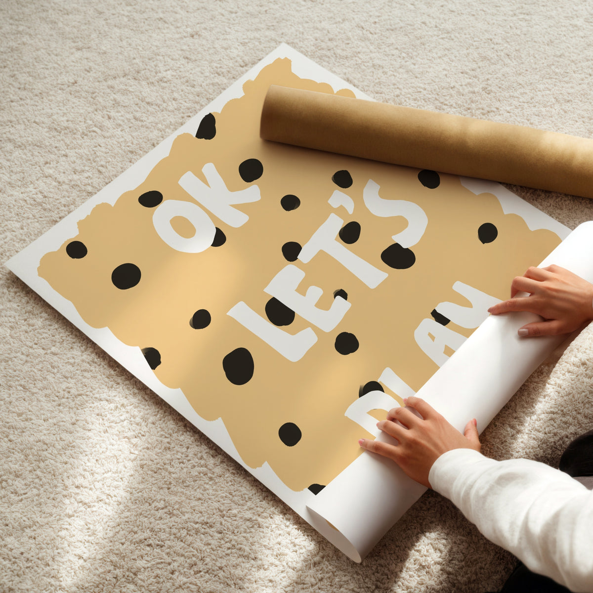 A person unrolls a large cookie-themed wall decal or poster on a beige carpet, with a brown paper roll nearby. The design shows a tan cookie with black polka dots and white text reading 'OK LET'S PLAY'.