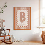 A framed personalized nursery print hanging on a white wall in a styled nursery room. The print features a large letter 'B' with the text 'is for Ballerina' on a striped background, enclosed in a light brown wooden frame.