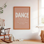 A framed wall art print with the word 'DANCE' in large white letters on a dusty pink background, hanging on a white wall in a softly styled room with a rocking chair, books, and a window.