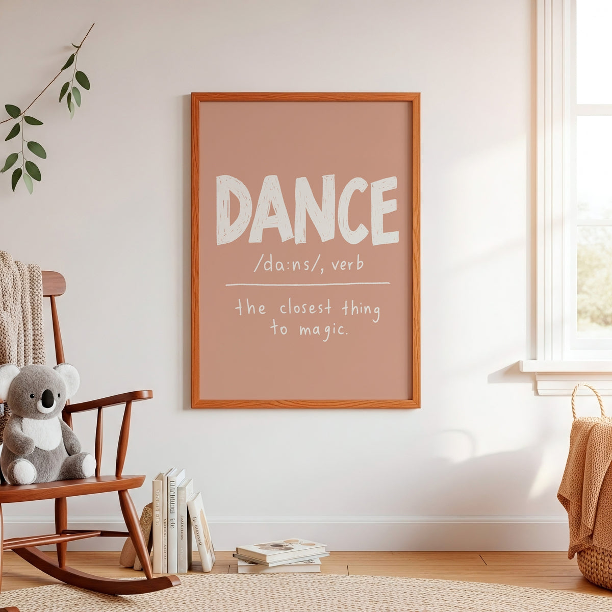 A framed wall art print with the word 'DANCE' in large white letters on a dusty pink background, hanging on a white wall in a softly styled room with a rocking chair, books, and a window.