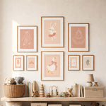 A styled nursery wall display featuring eight framed pastel art prints with ballet-themed illustrations, including a bunny, ballerina tutus, duck, and decorative monograms, arranged above a wooden shelf with toys and books.