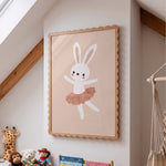 A framed children's wall art print featuring a white bunny in a pink tutu on a warm beige background, hanging on a white wall in a styled nursery with wooden beams, toys, and a macrame hanging visible below.