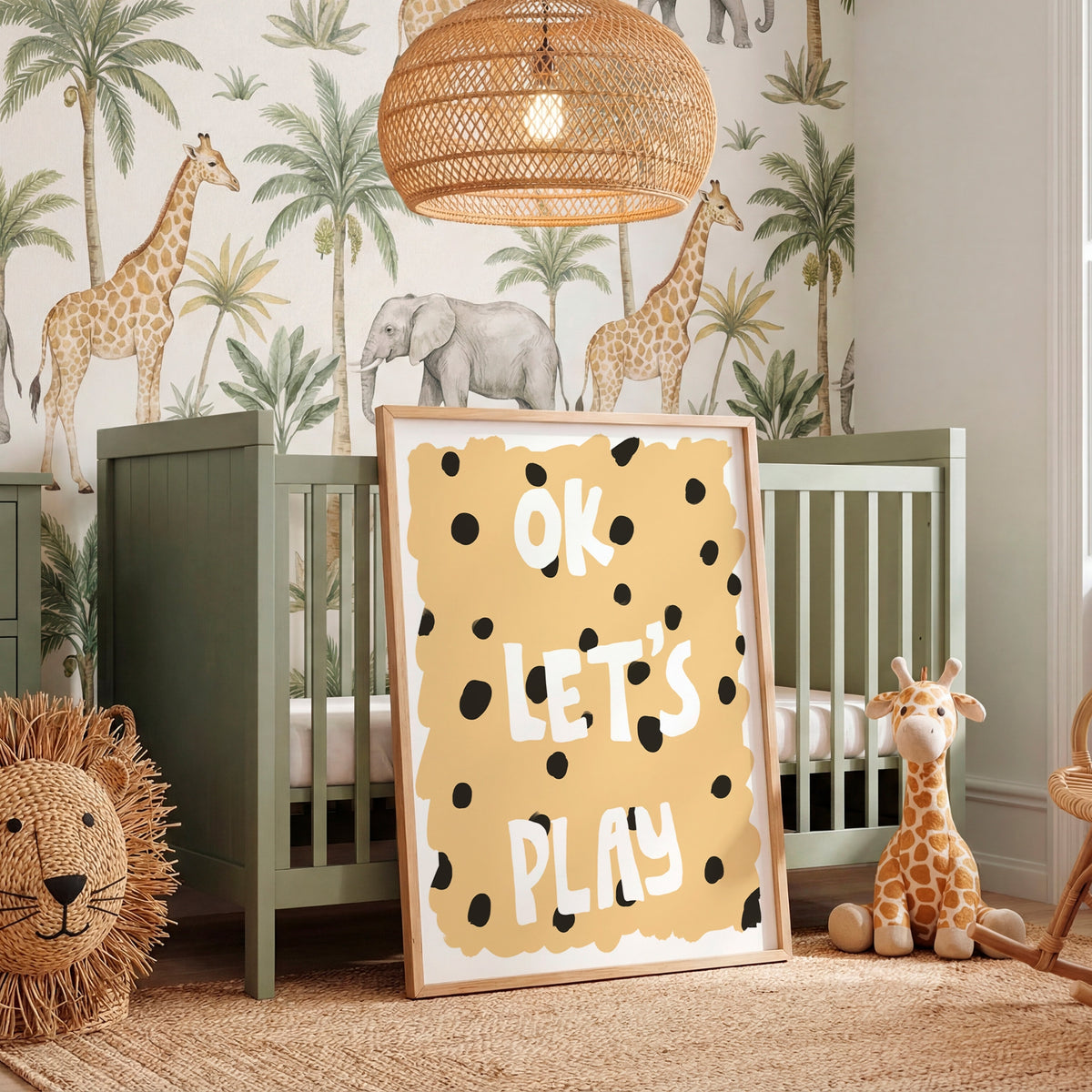 A styled nursery room featuring a sage green wooden crib, a large framed 'OK LET'S PLAY' sign with black polka dots on a yellow background, safari-themed wallpaper with giraffes and palm trees, a woven rattan pendant light, and a plush giraffe toy.