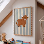 A framed wall art print featuring a cartoon Highland cow on vertical blue-gray and cream stripes, displayed in a child's bedroom with toys, a macrame hanging, and wooden beams visible.