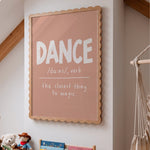 A framed typographic wall art print hanging on a light wall in a styled room. The print has a dusty pink background with the word “DANCE” in large white letters and a definition underneath, surrounded by a scalloped natural wood frame.