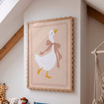 A framed wall art print featuring a white goose wearing a pink bow, hanging on a beige wall in a softly styled room with wood beams and small plush toys visible below.