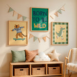 Three framed children's wall art prints hang above a wooden bench in a styled nursery. The center print says 'BORN TO BE WILD' on a green background, the left print shows a cartoon tiger on a skateboard, and the right print shows a green dinosaur on a plaid background.