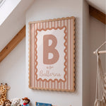Personalized nursery wall art featuring a wooden frame with pink and white vertical stripes, a large letter B, and the text 'is for Ballerina' in a nursery room setting.