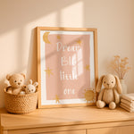 A framed nursery wall art print reading “Dream BIG little one” in white script with gold stars, a crescent moon, and a sun on a blush pink background, styled on a wooden dresser with plush toys and dried flowers in a warm, sunlit room.