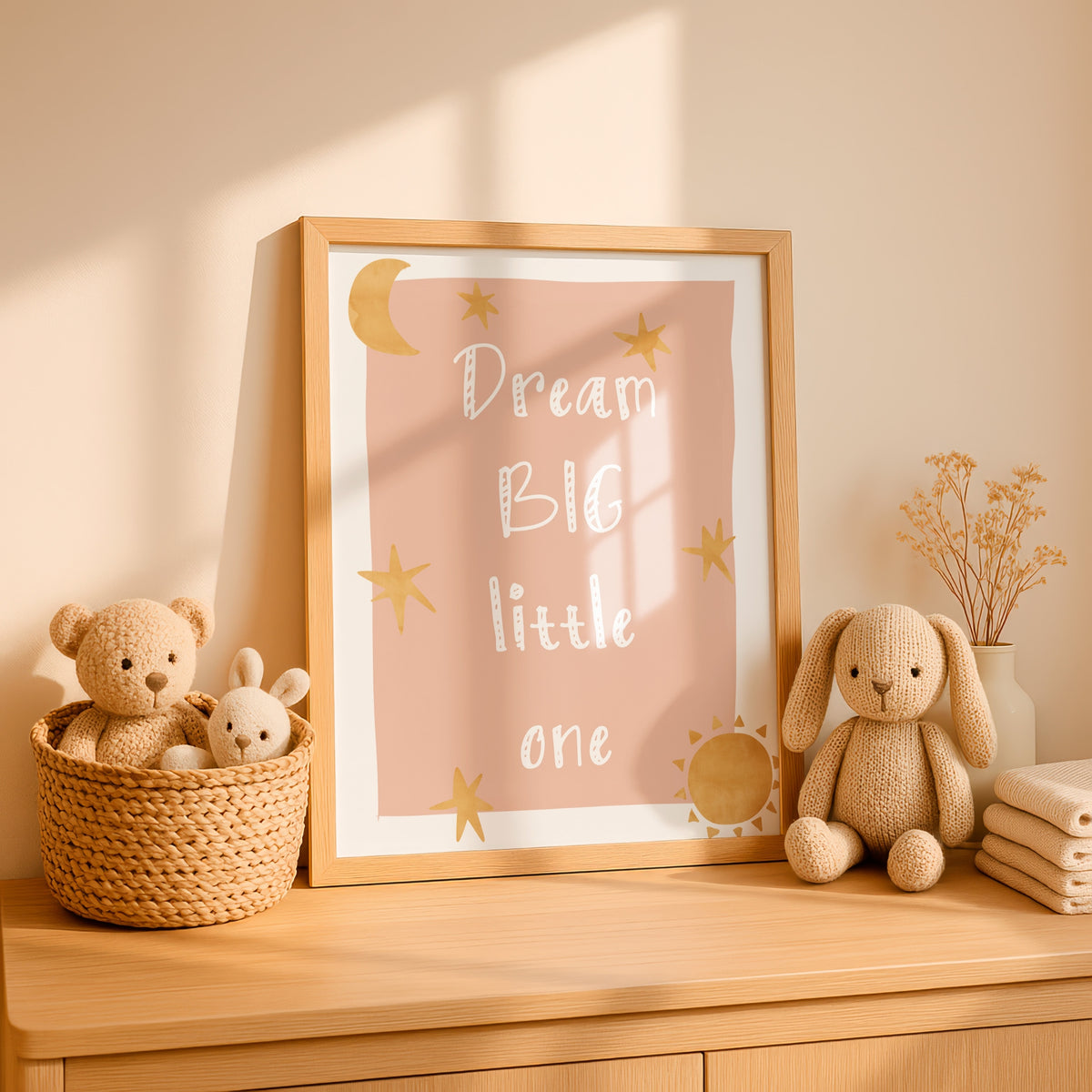A framed nursery wall art print reading “Dream BIG little one” in white script with gold stars, a crescent moon, and a sun on a blush pink background, styled on a wooden dresser with plush toys and dried flowers in a warm, sunlit room.