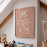 A framed wall art print featuring white line art of ballet pointe shoes on a dusty pink background, styled in a child's bedroom with toys and a macrame hanger.
