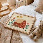 A framed art print featuring a red barn, yellow flowers, and red tulips, styled on a white bed next to a teddy bear and a knit blanket.