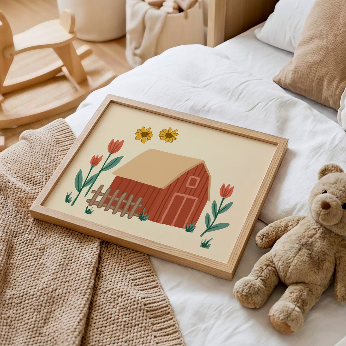 A framed art print featuring a red barn, yellow flowers, and red tulips, styled on a white bed next to a teddy bear and a knit blanket.