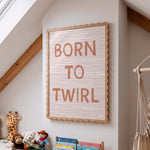 A framed letter board sign with the words 'BORN TO TWIRL' in dusty rose lettering on a white striped background, hanging on a light gray wall in a child's room.