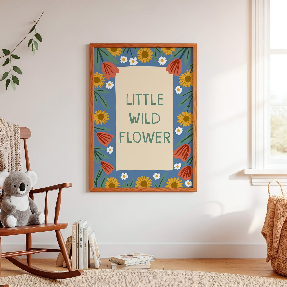 A framed wall art print reading “LITTLE WILD FLOWER” hangs on a white wall in a softly styled nursery. The print has a blue border with yellow sunflowers, white and orange flowers, and green leaves, and is shown in a room with a wooden rocking chair, books, and a window.