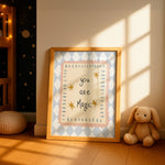 A framed nursery art print leaning against a wall in a sunlit room. The print has a light blue and white diamond pattern border, a pale pink inner border, and the words 'you are Magic' in black script with small gold stars.