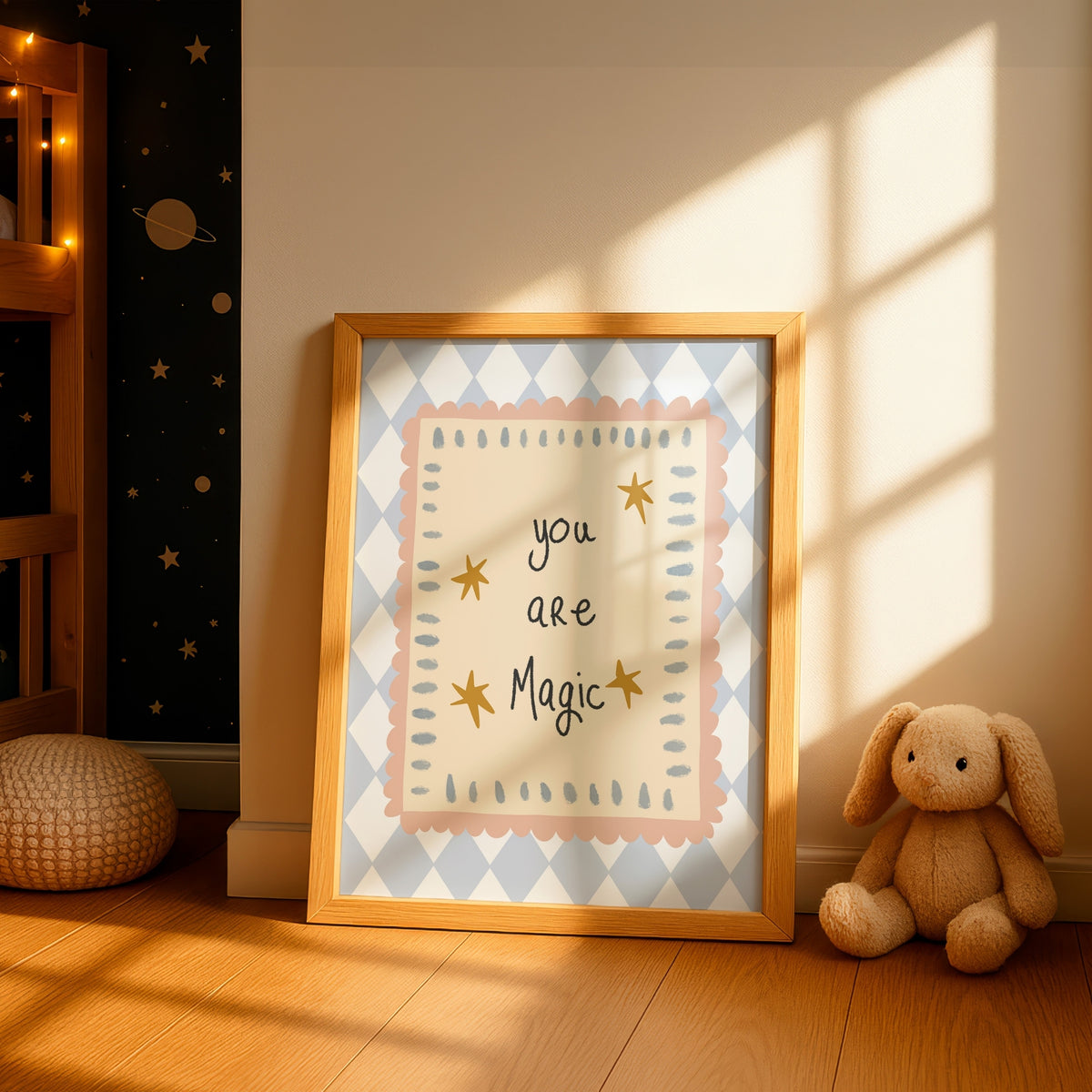 A framed nursery art print leaning against a wall in a sunlit room. The print has a light blue and white diamond pattern border, a pale pink inner border, and the words 'you are Magic' in black script with small gold stars.
