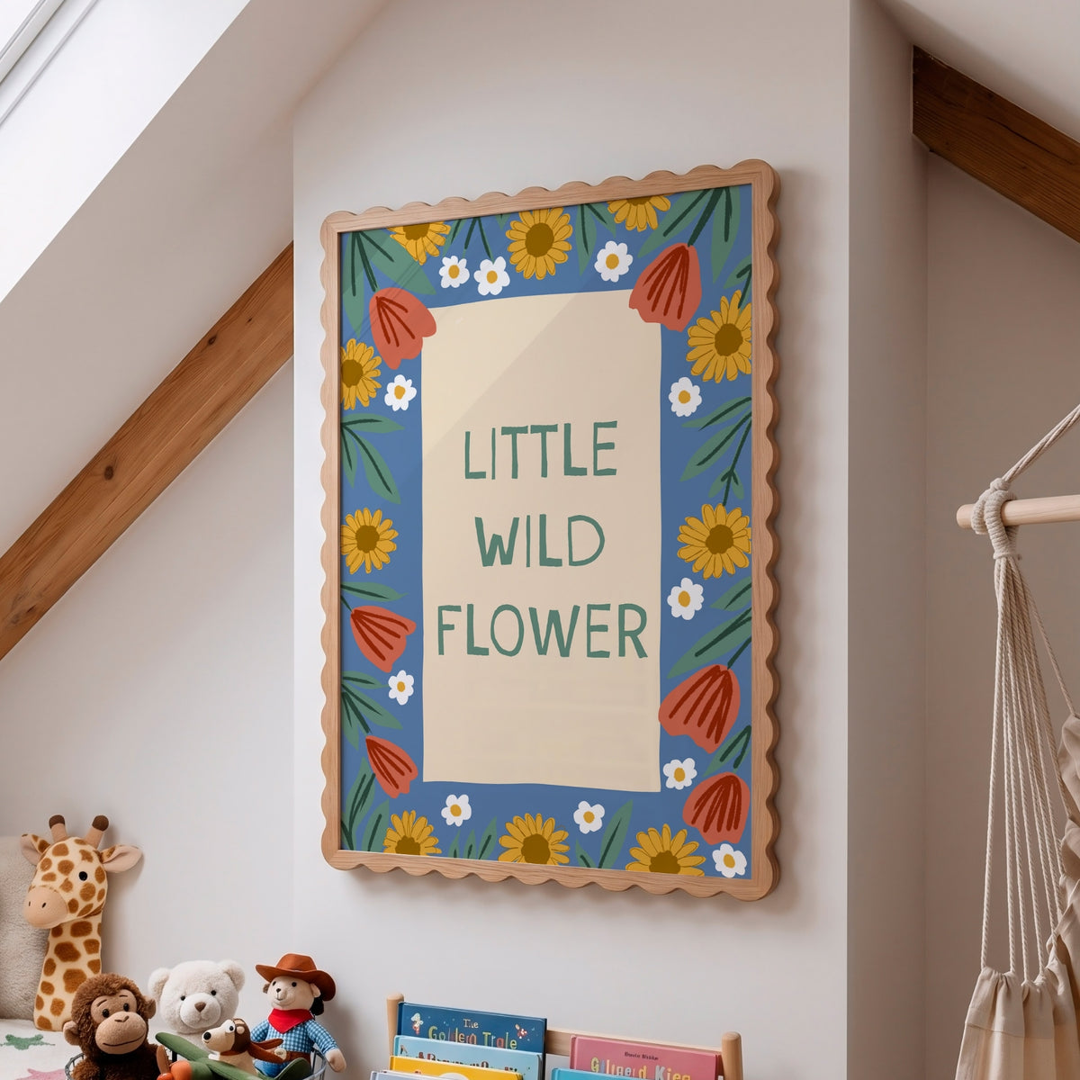 A framed wall art print with the words “LITTLE WILD FLOWER” in green lettering on a cream center panel, surrounded by a blue border with yellow sunflowers, white daisies, orange flower shapes, and green leaves. The frame hangs on a white wall in a child’s room with toys, books, and a macrame hanging visible.