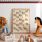 A framed alphabet nursery art print displayed on a wall in a styled children's playroom. The print features colorful illustrated letters and words with seasonal and everyday objects.