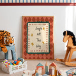 A framed nursery wall art print leaning against a white wall in a styled children's room. The print features the text 'Jingle All The Way' with gold stars on a cream background, bordered by green and terracotta patterns.