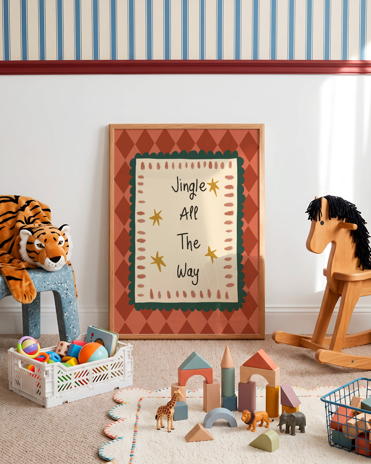 A framed nursery wall art print leaning against a white wall in a styled children's room. The print features the text 'Jingle All The Way' with gold stars on a cream background, bordered by green and terracotta patterns.