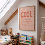A styled children's bedroom corner with a slanted ceiling, skylight, wooden beams, a bed with stuffed toys, a small wooden book rack, and a framed wall print featuring the word 'COOL' in orange-red lettering on a pale pink background.