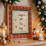 A framed Christmas art print with the text 'Jingle All The Way' displayed on a wooden mantel, surrounded by holiday decorations including a lit lantern, nutcracker, garland, and ornaments.