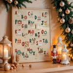 A framed Christmas alphabet art print displayed on a wooden mantel, surrounded by warm holiday decor including a lantern, nutcracker, pine garland with gold ornaments, and glowing candles.
