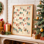 A framed Christmas alphabet art print leaning on a fireplace mantel, surrounded by wrapped gift boxes, a pinecone, evergreen branches, and a decorated Christmas tree.