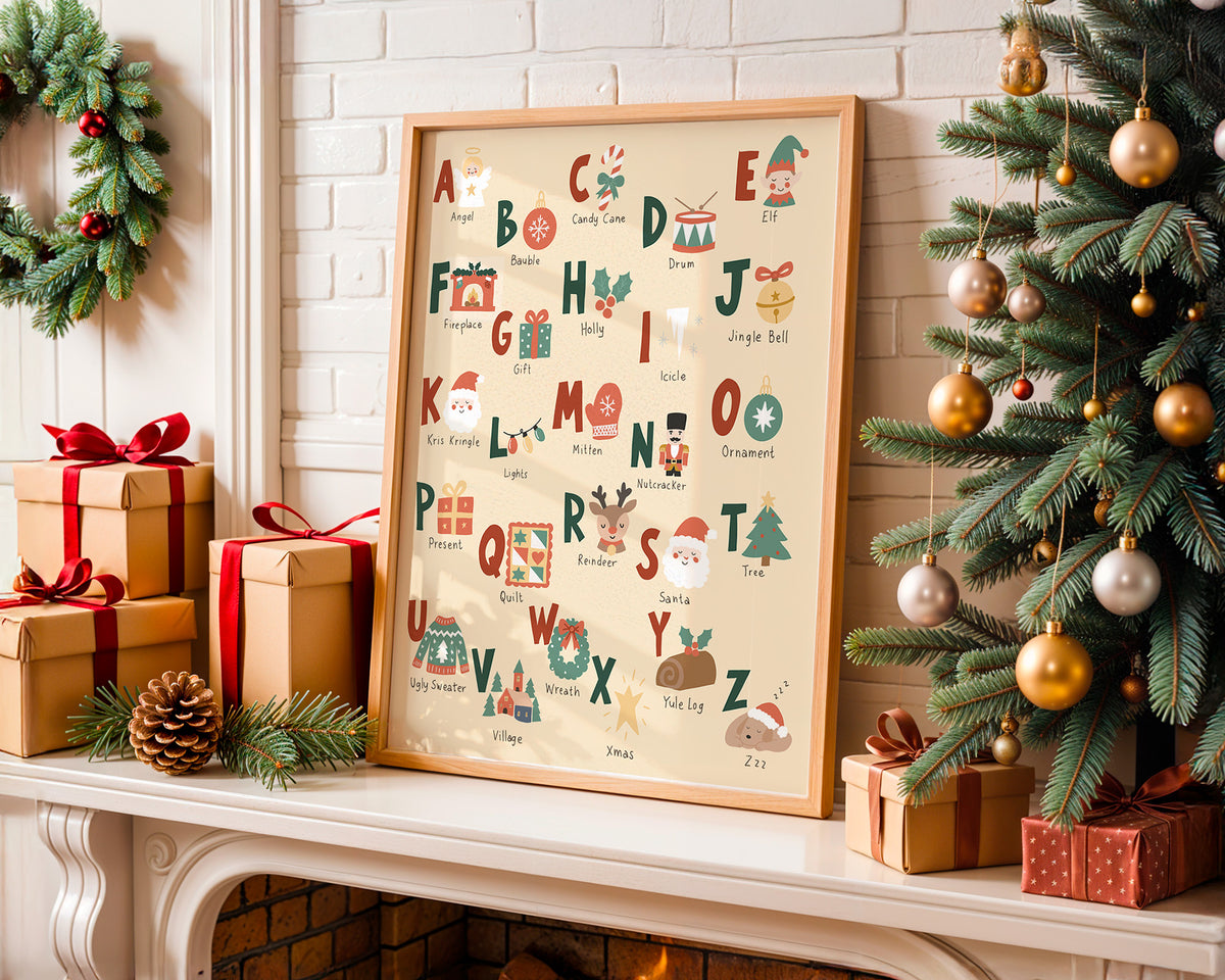 A framed Christmas alphabet art print leaning on a fireplace mantel, surrounded by wrapped gift boxes, a pinecone, evergreen branches, and a decorated Christmas tree.