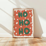 Framed wall art leaning against a white wall on a light wood floor, featuring the words “HO HO HO” in green on a terracotta background with white polka dots.