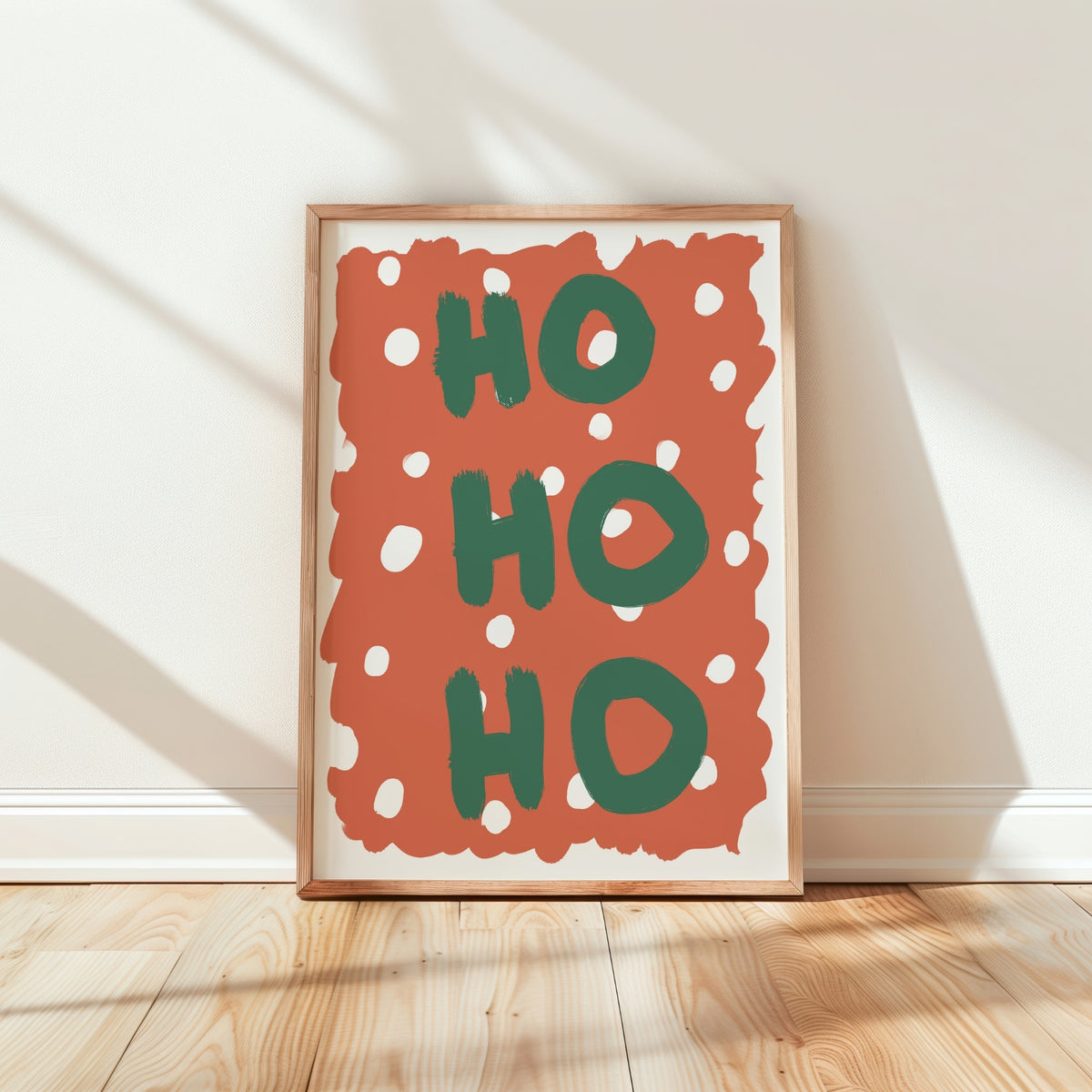 Framed wall art leaning against a white wall on a light wood floor, featuring the words “HO HO HO” in green on a terracotta background with white polka dots.