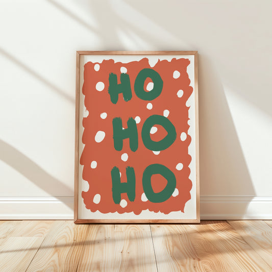 Framed wall art leaning against a white wall on a light wood floor, featuring the words “HO HO HO” in green on a terracotta background with white polka dots.