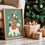 A framed Christmas art print leaning against a beige sofa on a wooden floor, next to wrapped gifts and a decorated Christmas tree. The print shows a white polar bear carrying a reindeer on its back, with a penguin riding the reindeer and a Santa figure at the top, all on a teal background with gold stars.