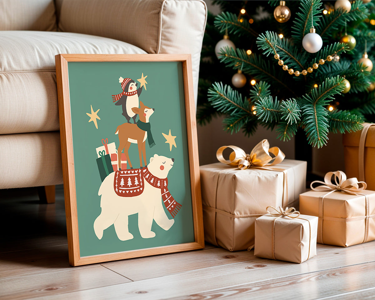 A framed Christmas art print leaning against a beige sofa on a wooden floor, next to wrapped gifts and a decorated Christmas tree. The print shows a white polar bear carrying a reindeer on its back, with a penguin riding the reindeer and a Santa figure at the top, all on a teal background with gold stars.